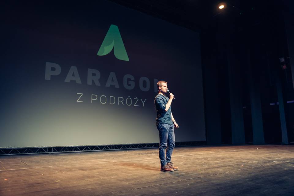Patryk Świątek z mikrofonem na scenie, na tle napisu Paragon z Podróży